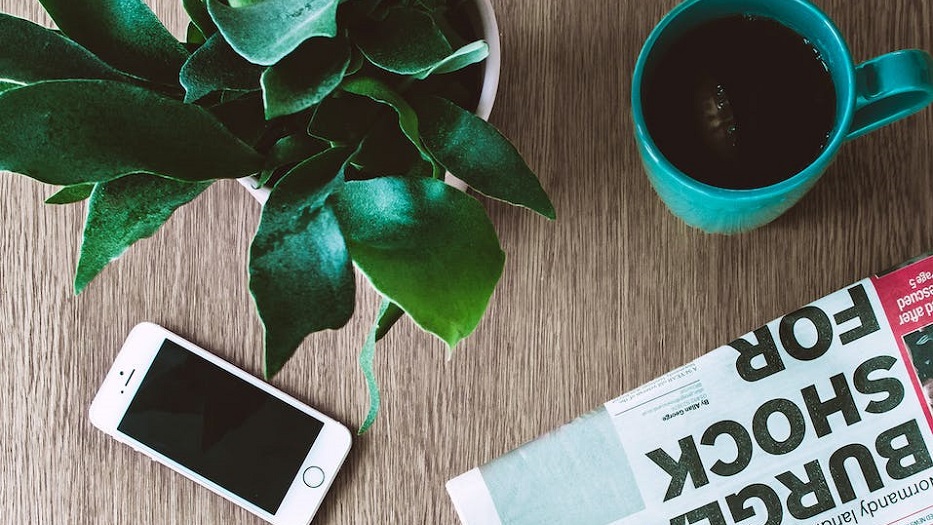 Symbolbild - Ein Mobiltelefon, eine Zeitung, eine Pflanze und eine Tasse stehen auf einem Tisch.
